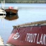 Words Trout Lake painted on row boat that is on shore with lake in background.