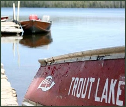 Words Trout Lake painted on row boat that is on shore with lake in background.