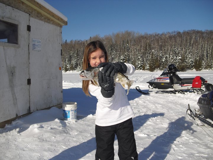 Young girl holding trout on frozen lake with ice fishing house and snowmobile in backgroun